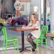 A woman with blonde hair wearing a brown dress and black tights and shoes sits at a table a table alone by a window